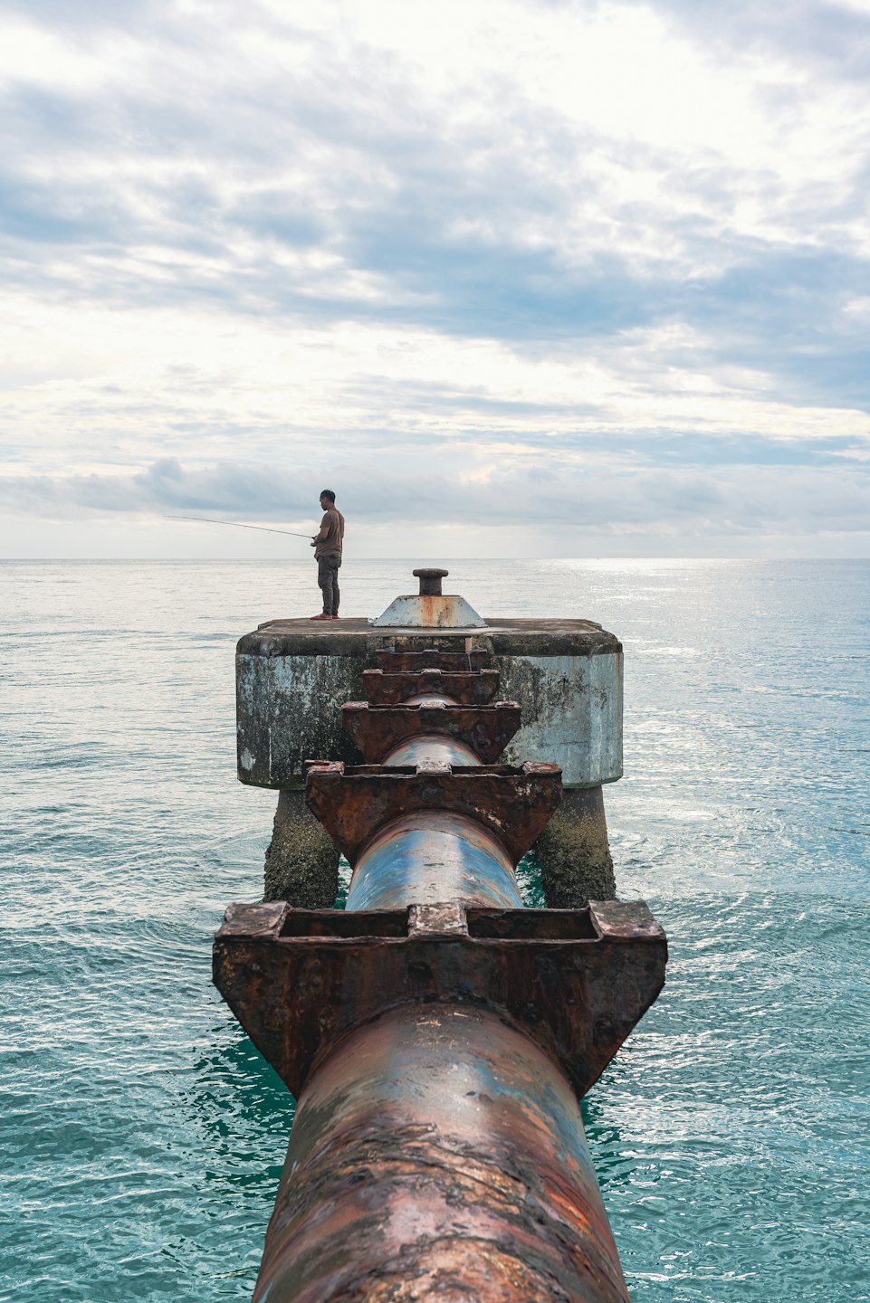 Mann fischt von Betonpier mit rostigen Rohren ins Meer.